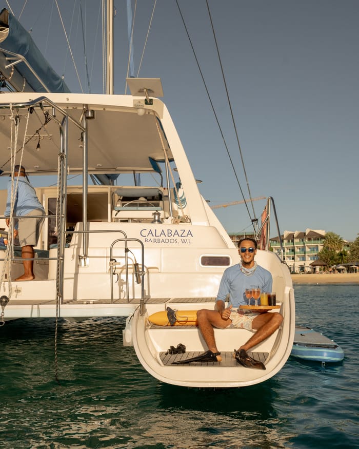 A crew member waits on Calabaza’s stern with a tray of drinks, the boat moored off the beach